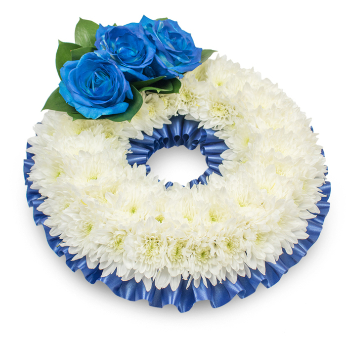 A traditional circular memorial wreath photographed from just above, set against a pristine white backdrop so the shadow beneath reads like a soft suggestion of weight and presence. The body of the wreath is densely composed of white chrysanthemums, their layered, papery petals creating a full, cushiony ring whose subtle doming and intricate surfaces invite a moment of close looking; the tiny, creamy-tinged centers add a whisper of warmth to the snowy mass. Placed with careful restraint at the upper left, three deep electric-blue roses rise from a bed of dark green leaves, their velvety petals unfurling in perfect spirals that offer a vivid, contemplative contrast to the white. Both inner and outer edges of the chrysanthemum band are bordered with a pleated cobalt satin ribbon, its glossy folds catching light and shadow to frame the floral work with refined dignity. The arrangement blends the traditional language of white flowers for remembrance with the uncommon, calming presence of blue - a palette suited to quiet reflection at wakes, memorial services or church gatherings across Brent Cross, Cricklewood and Golders Green. You can almost sense the subtle, dry fragrance of the chrysanthemums and the cool touch of satin ribbon; every element-flower choice, ribbon pleat, foliage placement-feels chosen to offer solace and a lasting memory in moments of farewell.
