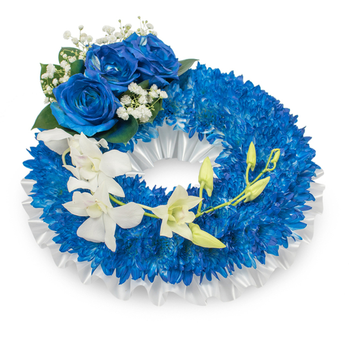 Presented against a clean white backdrop, this circular sympathy wreath is crafted from a dense carpet of small, luminous deep-blue chrysanthemums that form a plush, voluminous ring. The chrysanthemums' clustered petals produce an almost textile-like surface-tight, layered and visually soothing-whose royal indigo tones convey calm and solemnity. A pleated white satin ribbon lines the wreath's inner opening, its soft ruffles creating a precise, satin frame that both cushions and highlights the vivid blue. At the upper-left curve, a cluster of three expansive blue roses commands attention; their velveteen petals open in a deeper sapphire, rich with shadow and subtle highlights, and are accented by airy sprays of tiny white baby's breath and a scattering of dark green leaves. From the mid-left the eye follows a gentle cascade of pristine white orchids sweeping toward the lower right-orchid blooms with refined, sculptural petals that seem almost translucent in cool light. Intermingled with these blooms are several light-green orchid buds at differing stages of unfurling, symbolising renewal and continuity amid remembrance. The arrangement balances contrasting textures-spiky chrysanthemums, plush roses, and silk-smooth orchids-while maintaining a composed, respectful mood. You can imagine it placed at a funeral chapel near Brent Cross Shopping Centre or set beside a memorial wreath stand on Hendon Way, where its palette of profound blues and pure whites offers a poignant tribute: serene, respectful, and tenderly made by practised hands.