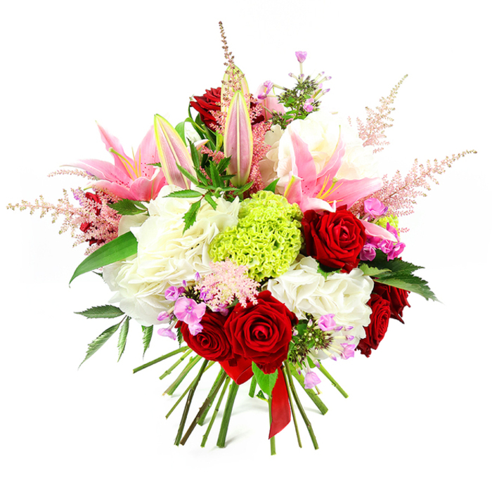 A richly detailed hand-tied bouquet sits against a pure white backdrop, its colours and textures showcased with striking clarity. The arrangement is composed in a rounded, full shape, brimming with blooms that create a sense of indulgent abundance, as though arranged for a dinner celebration in one of the family homes surrounding Brent Cross. Deep crimson roses take centre stage, their velvety petals folded in intricate spirals, symbolising love and elegance. Nestled among them, generous pink lilies open into broad, star-like flowers, each petal smooth and slightly curved, leading the eye toward pale, delicately speckled throats and fine stamens lightly dusted with pollen. A few lilies remain closed or only just beginning to open, their long buds introducing height and a quiet sense of expectation. Filling the spaces around them, clusters of soft white flowers-similar to large, ruffled carnations or hydrangea-create a luxurious, cloud-like foundation that supports the more structured blooms above. At the centre of this softness, a vivid lime-green sphere of finely textured florets, almost certainly Green Trick dianthus, offers an unexpected contemporary accent, with a tactile, mossy appearance that contrasts beautifully with the smooth petals surrounding it. Slender plumes of pale pink astilbe rise gently from within the bouquet, their feathery tufts giving the design lightness and movement. Delicate fuchsia and lavender filler flowers peek through, adding tiny bursts of jewel-like colour. Dark, glossy green leaves weave discreetly among the blossoms, providing depth and a fresh, garden-like feel. At the base, the natural stems are drawn together in a visible bundle and tied with a deep red satin ribbon, highlighting the hand-crafted nature of the piece and the skill typical of Brent Cross Flowers. The bouquet conveys warmth, romance, and celebration, perfect for anniversaries, birthdays, or thoughtful deliveries across Brent Cross and the surrounding North West London communities.