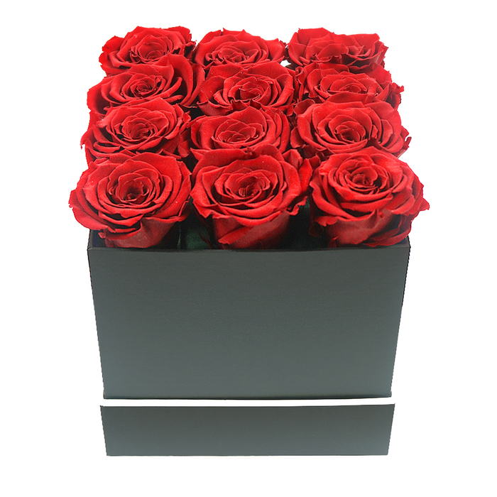 A striking floral composition dominates this close-up photograph: twelve preserved red roses arranged with exacting precision in a square, matte black gift box, ideal for a romantic gesture anywhere around Brent Cross and nearby Golders Green. The roses, all uniform in size and fullness, are lined up in three neat rows of four, completely filling the top of the box so that only the blooms, and not the stems, are visible. Each flower is in full, open bloom, revealing layer upon layer of velvety petals. The red tones range gently from deep, almost garnet shadows in the centres to slightly brighter crimson at the curled outer edges, creating a soft play of light and dark across the surface of the arrangement. The petals appear smooth and plush, suggesting the long-lasting quality of high-end preserved roses. The box that holds them has a smooth, matte finish and sharply defined corners, its deep black tone acting as a dramatic, but understated, frame. Around the lower part of the box, a fine, bright white band circles the container, lending a clean, graphic line that emphasises the modern design. The entire arrangement is photographed against a spotless white background, with soft, diffused lighting that illuminates every curve and fold without casting harsh shadows. Only gentle shading beneath the petals and along the edges of the box hints at depth and volume. The impression is of a gift that combines minimalism with opulence, suitable for a Brent Cross couple celebrating an anniversary, a Valentine's Day surprise delivered to a flat near the station, or a chic decorative piece in a contemporary living room. It feels like an expression of enduring love and thoughtful taste, designed to remain beautiful for months rather than days.