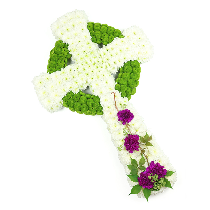 In this high-quality studio image, a Celtic cross floral tribute crafted by Brent Cross Flowers is displayed in full, resting on a spotless white background that draws attention to every carefully placed bloom. The cross is composed primarily of crisp white chrysanthemums, arranged so densely that they form a continuous, cushioned surface along the vertical stem and the outstretched horizontal arms. The slightly domed placement of each flower head creates a sense of depth and softness, as if the entire form has been gently upholstered in petals. Threaded through this white canvas is the ancient Celtic cross design, rendered in bright lime green button chrysanthemums. These neat, rounded blooms form a prominent ring at the centre where the arms meet and continue in narrow, interwoven bands that trace along the arms and stem, echoing the intricate knotwork often associated with Celtic spirituality and heritage. This combination of white and green is both serene and fresh, evoking themes of purity, renewal, and the continuity of life, sentiments that resonate deeply at funeral and memorial services across Brent Cross, Cricklewood, and the surrounding North London area. On the lower section of the vertical arm, three striking clusters of deep purple carnations are arranged to create a strong focal accent. Their lush, ruffled petals and intense colour range from royal purple to warm magenta, lending the tribute an emotional richness that balances the calm of the white chrysanthemums. Around these carnation groups, short sprigs of glossy green foliage and a few fine, curling woody stems add natural texture and a sense of gentle movement without disturbing the overall neatness of the design. The lighting is bright but soft, enhancing the vibrancy of the colours and the freshness of the flowers. The resulting impression is one of quiet elegance and heartfelt remembrance, a meaningful floral cross for honouring a loved one with both faith and grace.