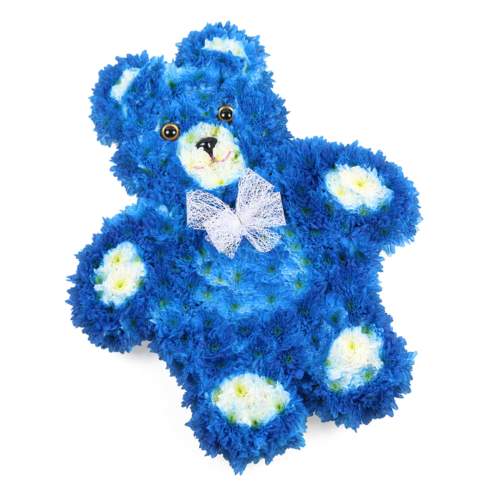 Against a bright white backdrop, a three-dimensional teddy bear crafted entirely from flowers becomes the poignant focal point of this image. The bear's body is generously shaped, with rounded limbs and a softly curved tummy all cloaked in tightly packed royal blue blooms. These small, multi-petalled flowers are arranged so densely that they create a smooth, plush-like surface, with subtle shadows deepening the blue and adding a sense of depth and form. To bring out the bear's features, the florist has used clusters of creamy white flowers, tinged with pale yellow at their centres, carefully set into the inner ears, rounded snout, and oval paw pads, mirroring the details of a classic children's teddy. The face holds a gentle, comforting expression: two round, glistening brown eyes with dark pupils, a neat black triangular nose placed at the centre of the muzzle, and a fine curved black line suggesting a stitched mouth. Around the neck is tied a delicate white mesh bow, its transparent folds softening the transition between head and body and adding a subtle sense of occasion. The bear lies flat but is photographed on a slight angle, giving the composition a quiet dynamism and making the tribute feel more like a cherished toy placed with intention. In Brent Cross and the surrounding North West London neighbourhoods, such a floral bear would be a deeply touching choice for a child's funeral, a baby boy's farewell, or a remembrance service at a local chapel, offering a visual language of innocence, peace, and the tender care of a skilled florist who understands the sensitive nature of these moments.