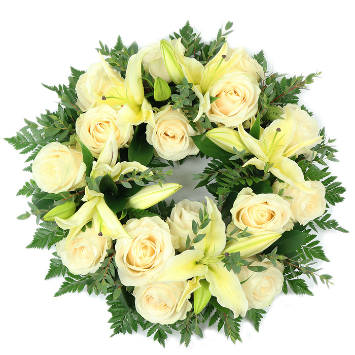 A meticulously assembled circular wreath photographed from above, blending cream-coloured roses and pale yellow lilies into a dense, elegant memorial piece-crafted in the spirit of Brent Cross floral tradition and well suited to services in the local area around Brent Cross Shopping Centre, Cricklewood, or Golders Green. The arrangement features roughly fifteen softly unfurling roses, their ivory petals layered in tight spirals that glow against the green underlayer. Scattered between them, lemon-tinted trumpet lilies provide taller accents; some open to reveal delicate stamens, others remain slender buds for layered texture and visual rhythm. The greenery is rich and mixed: fine, feathery fern fronds create a soft halo along the wreath's edge while rounded, grey-green sprigs-resembling eucalyptus-sit lower, offering shadowed depth and a faint, fresh aroma. Set flat on a clean white background, the wreath's uninterrupted circle speaks of continuity and remembrance, its symmetry conveying dignity and calm. Soft, natural light reveals subtle variations in petal tone and the delicate veins of the foliage, enhancing the sense of careful handwork. Intended as a sympathetic tribute for funerals, memorial services, or heartfelt condolences delivered across Brent Cross and neighbouring streets, this piece balances formality with tender reassurance-an understated, respectful expression of remembrance shaped by local floristry expertise.