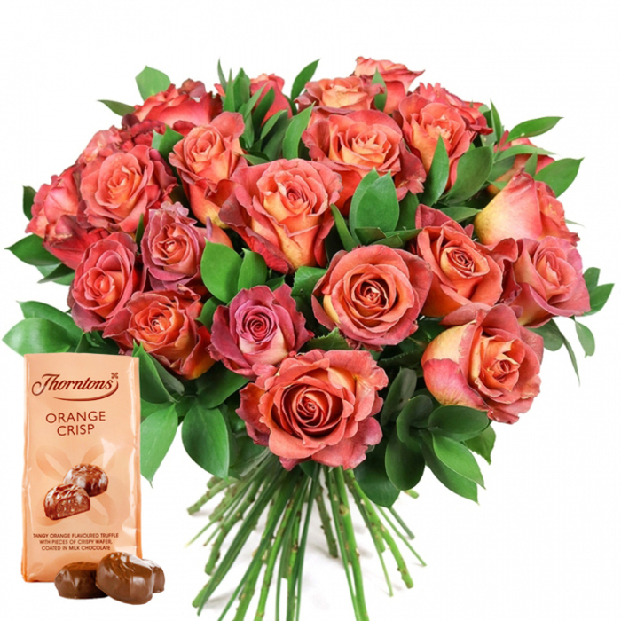At the centre of this tastefully arranged image stands a magnificent bouquet of fresh roses, masterfully crafted by Brent Cross Flowers and set against a spotless white backdrop. Each rose exhibits a gentle gradation of warm hues: tight yellow and creamful centres bloom into rounded petals glowing with shades of peach, coral, and a deeper, rosy pink, producing a mesmerizing bi-colour display. The velvety petals nestle densely together, giving the bouquet an opulent fullness reminiscent of the grand gardens near Brent Cross Station. Interspersed amongst the blossoms, glossy greenery-possibly ruscus or eucalyptus-adds a crisp, verdant element that complements the warmth of the roses. Arranged in the lower-left foreground, the iconic Thorntons Orange Crisp chocolate pouch stands proud, its light cream packaging displaying the Thorntons logo and the ORANGE CRISP label, complete with a graphic of glossy chocolate truffles. Below this, the pouch's description tempts with 'tangy orange flavoured truffle with pieces of crispy wafer, coated in milk chocolate.' Two real truffles rest close by, shining with a lacquered finish. The bouquet's neat green stems peek from the base, reinvoking the image of freshly cut flowers delivered straight from a Brent Cross workshop. The entire arrangement, in focus against a spotless background, makes a perfect, thoughtful gift for any special event-from anniversaries in Temple Fortune to birthdays near Gladstone Park-embodying the love and care synonymous with this local florist.