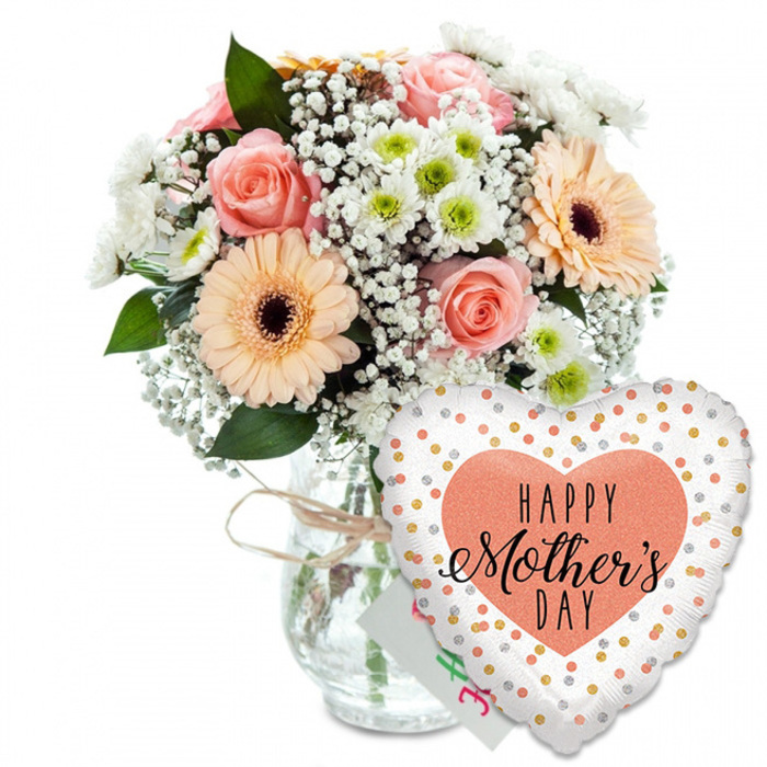 Photographed against a bright, uncluttered white background, this Mother's Day ensemble feels both modern and heartfelt, the sort of gift Brent Cross Flowers might prepare for a doorstep surprise on a quiet residential street nearby. On the left side of the frame stands a clear glass jar-style vase, its simple, unembellished shape chosen to let the flowers shine. Around the top, a piece of pale brown raffia or twine is tied in a small bow, adding a touch of rustic warmth that recalls handmade gifts and family traditions. The vase brims with a lush, rounded bouquet of mixed seasonal blooms in soft, flattering pastels. Several peach and light pink roses open fully at the centre, their layered petals spiralling outward in gentle curves that suggest affection and gratitude. Complementing them are prominent peach-toned gerbera daisies, their symmetrical petals radiating from dark brown centres that provide a striking visual anchor. Between these star blooms, airy clusters of white baby's breath are threaded delicately, creating a light, cloud-like texture. Smaller daisy-like white chrysanthemums with faint apple-green centres fill the spaces, lending extra volume and a fresh, lively accent reminiscent of springtime walks along the nearby green spaces by the North Circular. Rich greenery in mixed tones of green surrounds and supports the flowers, with leaves that peek between petals, adding depth, contrast, and the suggestion of natural movement. Resting just behind the bouquet is a small white greeting card, its printed colours only partly visible, hinting at a private message from child to mother. To the lower right, overlapping the vase, a large heart-shaped foil balloon takes pride of place. Its surface is bright white, scattered with tiny metallic gold and silver dots that look like floating confetti, giving a playful, celebratory lift. At the centre is a solid peach or coral heart, within which the message HAPPY Mother's DAY appears in bold black letters; the word HAPPY is written in a simple uppercase font, while Mother's DAY is rendered in a more flowing, script-like style that feels softened and personal. Together, the bouquet, card, and balloon form a thoughtfully coordinated Mother's Day gift, capturing the warmth, joy, and appreciation shared in homes around Brent Cross.
