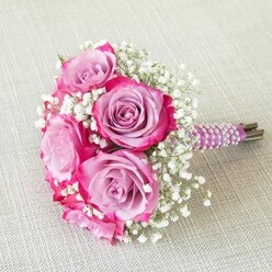 This captivating bouquet shines with the subtle artistry that defines weddings and celebrations throughout Brent Cross, where tradition and modern flair come together. At the heart of the arrangement, velvety roses in soft lavender and radiant fuchsia hues create an alluring two-tone gradient, their petals gently opening to reveal a sense of unfolding joy. The texture of the roses contrasts beautifully with the cloud-like clusters of white baby's breath, whose tiny blossoms seem to dance amid the larger blooms. The bouquet is expertly assembled, its natural stems wrapped with a rich purple satin ribbon, smoothly adorned with spirals of shimmering pearls-a tactile and visual detail reminiscent of the refined décor seen at local Synagogues or reception halls near Hendon Way. Beneath the elegant wrapping, the bouquet's green and brown stems remain visible, adding a touch of rustic charm. Soft daylight spills across an off-white, linen-textured surface, enhancing the freshness and colour of each flower, much like morning sunlight brightening local markets near Prince Charles Drive. With every detail considered, this arrangement radiates both intimacy and sophistication-an ideal statement piece for bridesmaids or an honoured guest, capturing the heartfelt spirit of Brent Cross occasions.
