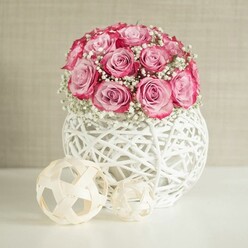 A carefully styled tabletop display shows a lush dome of pink roses and white baby's breath emerging from a white, basket-like vase, radiating a sense of gentle celebration that would suit a cosy living room off Hendon Way in Brent Cross. The bouquet itself is compact and full, formed from numerous roses gathered closely so the blooms touch and overlap. Each rose reveals a gradient of colour, with soft, powdery pink at the center deepening into more saturated, almost fuchsia tones along the outer petals, giving the arrangement depth and movement. Between the roses, clouds of delicate gypsophila weave in and out, their tiny white flowers and fine stems adding a feathery, cloud-like texture that lightens the whole design. Imagined as freshly arranged, the flowers feel velvety and cool to the touch, carrying a subtle, classic rose fragrance that suggests affection and thoughtfulness. They sit in a spherical white vase crafted from interwoven strands that resemble white-painted wicker or rattan, the open, lattice-style structure casting faint shadows onto the white surface beneath. To the left of the vase, two decorative spheres made from pale, natural-looking wooden strips echo the woven texture, their open, cage-like forms adding a minimalist, organic touch. The larger sphere sits slightly behind the smaller one, creating a gentle sense of depth. The background is a warm, neutral beige wall with a soft, slightly mottled texture that keeps the focus firmly on the flowers. Bright but diffused lighting, similar to natural daylight filtering through a north London window, illuminates the arrangement evenly, highlighting every petal and strand. Overall, the scene suggests a refined, modern floral centerpiece suited to marking a quiet anniversary dinner, a Mother's Day breakfast, or a thoughtful same-day delivery surprise in the Brent Cross area.