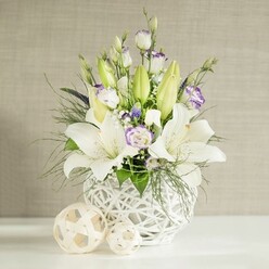 An elegant, contemporary floral design ideal for a wedding table is captured here, glowing softly in natural light. The arrangement is gathered in a unique round white vase with an open, basket-like structure, its crisscrossing strands creating gentle shadows that echo handcrafted rattan work. Rising proudly from this base are several white lilies, some fully open with star-shaped petals stretched wide, their centers marked by slender stamens dusted with golden pollen, while others remain in pale green buds, giving the arrangement height and anticipation. Around the lilies, white lisianthus blossoms soften the composition, their petals layered like silk and brushed in places with hints of lavender and lilac, so subtle they appear almost watercolor. Tucked among these are tiny accents of deeper purple flowers, offering a quiet contrast that enhances the bridal palette without overwhelming it. Feathery green foliage and broader, lush leaves weave throughout, some arching over the rim of the vase to soften its outline and introduce a touch of natural wildness. On the smooth white surface below, two decorative spheres made from light, woven material are placed beside the vase, their rounded shapes and open patterns mirroring the container's design and lending a harmonious balance. The backdrop is a light beige wall with a fine horizontal texture that suggests a calm, well-curated interior. The overall impression is one of tranquillity and refined celebration, a centerpiece that could sit beautifully on reception tables at a Brent Cross wedding venue or at a family gathering near Golders Green, crafted to bring a whisper of fragrance, purity, and romance to local celebrations.