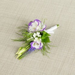 Set on a calm, light beige fabric that looks like finely woven linen, this boutonniere has the intimacy of something chosen with real care on a quiet morning in Brent Cross. Two lisianthus blooms anchor the design, their petals opening in creamy white layers with delicately ruffled edges washed in vibrant purple, as if they have been gently tinted by dusk light. Around them, tiny white filler flowers gather in soft clusters, adding a misty, romantic texture that feels almost like breath on a windowpane. Rising at one side, a vertical stem of deep purple muscari forms a line of tiny, bell-shaped buds, stacked closely together and rich in colour, creating a subtle contrast between structure and softness. Small, pale green buds nestle near the focal flowers, hinting at blossoms still to come and lending a sense of quiet anticipation that mirrors the emotions of a wedding morning. Wisps of fine, fern-like greenery frame the whole boutonniere, light and feathery, suggesting it might have been freshly picked from the greenery around local parks near Brent Cross. The stems are gathered cleanly and bound with a smooth band of white satin ribbon, tightly wrapped and finished in a modest bow, ready to be pinned to a lapel with ease. The overall mood is serene and thoughtful, as though intended for someone who prefers their flowers to speak in gentle tones-perfect for a groom, groomsman, or prom guest making their way to a ceremony or celebration in North West London's quieter residential streets.