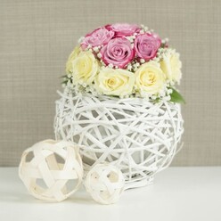 A romantic floral display sits at the centre of a minimalist white table, capturing the soft elegance often chosen for weddings and chic celebrations around Brent Cross. The arrangement is made up of about a dozen roses, gathered into a compact dome that rises from a white wicker globe vase. The roses themselves are a tender blend of colour: pastel pinks with hints of lavender and touches of deeper fuchsia mingle with buttery cream and ivory blooms. Their petals are velvety and fresh, some buds just beginning to unfurl while others are fully open, spiralling outward in layered rosettes. Sprigs of delicate white baby's breath are scattered thoughtfully between the roses, their tiny clustered blossoms lending a misty brightness that fills in any spaces and gives the bouquet a cloud-soft edge. Cradling the flowers is a spherical vase made from fine white-painted branches or wicker, interwoven to create a hollow, airy sphere. This open latticework allows light to slip through and reveals a glimpse of the stems and inner container, adding dimension and visual curiosity. On the left side of the same white surface are two additional decorative spheres crafted from pale natural rattan, with an open, woven structure that mirrors the design of the vase and contributes to the natural, handcrafted feel. Behind, a neutral beige wall with subtle texture serves as a calm backdrop, enhancing the gentle tones and clean lines. The scene feels ideal for a wedding breakfast near Brent Cross, a stylish engagement party, or a carefully curated dinner at home, where a floral centerpiece can quietly tie the room together without overwhelming it.