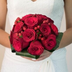 In this elegant depiction from Brent Cross, the lush, sophisticated bouquet stands out as a testament to traditional romance with a modern twist. Each deep crimson rose, meticulously selected for its fullness and luxurious texture, appears to swirl in perfect harmony at the centre of the bridal arrangement. Tucked between the roses, clusters of radiant red hypericum berries emerge, their playful forms adding lively punctuation to the otherwise classic display. Surrounding the blooms, wide, sculpted green leaves create a polished frame, evoking the verdant beauty of Brent Park while offering a crisp contrast to the vibrant reds. The bride's hands, with freshly manicured nails, gently cradle the rounded bouquet, emphasising its significance and the unity it represents. Her wedding gown, with intricate lacework and flowing fabric, adds an elegant backdrop that complements the passionate palette of the bouquet. The scene is softly illuminated, enhancing the bouquet's rich tones and inviting viewers to imagine the subtle scent and tactile allure of expertly arranged Brent Cross flowers. This bouquet offers a bold, heartfelt accent to weddings in the area-perfect for couples seeking both drama and grace on their special day.
