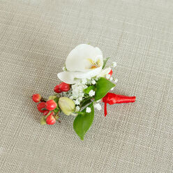 An artfully composed boutonniere is captured from above, lying on a pale oatmeal-coloured, woven fabric that looks like natural linen, the kind often used on elegant bridal tables in Brent Cross venues. In the centre, a flawless white Phalaenopsis orchid opens in a serene, sculptural shape, its broad petals smooth and slightly luminous against the neutral background. At the orchid's core, a subtle blend of creamy yellow and soft orange adds depth, drawing the eye inward like a quiet glow. Surrounding this focal bloom, sprays of delicate baby's breath form a fine, mist-like halo, each tiny white floret contributing to a sense of lightness and romance. To one side, a compact cluster of glossy, round red berries brings a decisive pop of colour, their rich crimson tones adding a hint of festivity that feels perfect for winter weddings or formal gatherings near Clitterhouse Playing Fields or events hosted around Brent Cross station. Intermingled with the berries are a few paler green buds, suggesting freshness and adding gentle variation in tone. Bright, smooth green leaves, both narrow and slightly broader, weave throughout the design, framing the white and red elements and reinforcing a feeling of natural abundance. The stems are tightly wrapped in a vivid red satin ribbon, tied into a neat, understated knot whose short trailing ends rest softly against the textured cloth. The lighting is gentle and even, allowing the contrasting surfaces-glossy berries, velvety petals, and woven fabric-to be clearly read. Overall, the corsage or buttonhole radiates refined celebration, perfectly suited to a groom, groomsman, or prom guest seeking a polished floral detail in the Brent Cross area.