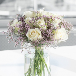 The photograph presents a tall glass vase cradling a lush bouquet of white roses and lavender-toned filler flowers, styled with the composed simplicity that suits a thoughtful sympathy arrangement in Brent Cross. The roses are the clear focus: around a dozen blooms, fully open and luxuriously layered, form soft spirals of petals in shades of white and cream. Their texture appears almost satin-like, with edges catching the light gently, giving each flower a subtle glow. Woven between them are delicate clusters of tiny flowers in pale lavender, soft lilac, and a hint of dusty pink, their fine, papery petals creating a misty veil that wraps gently around the roses. This combination of shapes and textures gives the bouquet a cloud-like fullness, while still feeling airy and refined. Beneath the blossoms, long green stems are visible through the transparent cylindrical vase, reaching down into crystal-clear water that enhances the sense of freshness, as though the arrangement might have just arrived from a local Brent Cross florist along Highfield Avenue or near the shopping centre. The vase rests on a clean, white surface, its base reflecting slightly, adding a delicate brightness to the lower edge of the scene. The background is softly out of focus, a wash of pale tones reminiscent of understated interiors in the residential streets off Hendon Way. Light falls softly from one side, casting gentle highlights and minimal shadows, contributing to the bouquet's calm and composed presence. The overall impression is one of quiet solace, a floral tribute designed not to shout but to speak gently, making it a fitting choice for memorial services, condolence visits, or sympathy deliveries across the Brent Cross area.