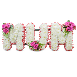 A deeply felt sympathy tribute presenting the word MUM in large, connected letterforms, each letter built almost entirely from small, snow-white chrysanthemums for a uniform, cloud-like surface. The white blooms are closely packed so that the letter faces appear soft and continuous, while a delicate ruffled ribbon in a muted pink frames each outer edge like a gentle embrace. A discreet dark green metal frame gives the standing display its shape, visible only where the letters meet, so the focus remains on the floral craftsmanship. Three vivid focal clusters enliven the design: a little posy of rich fuchsia-pink roses with lush ferny foliage and tiny coral hypericum berries nestles into the upper left of the first M; a mirrored cluster sits in the lower curve of the U; and the final M carries the third grouping at its top right. The contrast of velvety rose petals and glossy berries against the feathery chrysanthemum faces creates an intimate tactile interplay and a subtle lift of colour across the tribute. In soft morning or chapel light the whites glow with quiet dignity and the pink trim warms the composition, while the scent remains restrained and respectful. Prepared with care for funeral and memorial settings, this arrangement embodies purity, love and remembrance. Created locally and familiar to families arranging services in and around Brent Cross - including nearby Hendon and Golders Green - Brent Cross Flowers often crafts pieces like this to offer comfort and a graceful farewell at wakes, memorial services or graveside dedications.