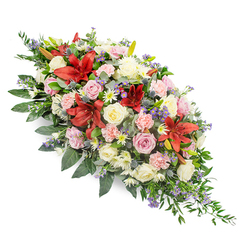 This image features an elegant double-ended casket spray, photographed on a pure white backdrop so the colours and shapes stand out with clarity and grace. The arrangement stretches horizontally, with its fullest point at the centre and both ends tapering to gentle points, making it suitable to rest along the top of a coffin at a farewell gathering in Brent Cross. Deep red Oriental lilies rise above the other flowers, their large, star-shaped blooms and prominent stamens adding drama and a sense of solemn beauty. Nestled among them are roses in shades of cream, pale pink, and ivory, some still partly closed, others fully open and layered, suggesting the unfolding of cherished memories. Frilled light pink carnations bring softness and volume, while clusters of white spray chrysanthemums with their many small petals add brightness without breaking the arrangement's calm and respectful tone. Threaded throughout are subtle touches of lavender and light purple filler stems, along with a few steel-blue thistle-like flowers that bring textural contrast and a cool note to balance the warmth of the reds and pinks. All the blooms are supported by a rich foundation of dark and mid-green foliage, including broad, waxy leaves and more delicate, ferny greenery that spills slightly beyond the flowers, creating a natural, flowing outline. The design feels thoughtful and composed, a floral expression of sympathy that would be at home in a chapel near Hendon Way or by the North Circular, crafted by a skilled local florist who understands how important it is for a final tribute to reflect both beauty and heartfelt emotion.