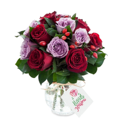 In this carefully balanced bouquet, deep, wine-coloured roses and soft lavender blooms converse in a gentle, measured rhythm: the crimson petals read passionate and plush, while the mauve roses bring a cool, calming counterpoint. The blooms are arranged densely into a rounded silhouette so that each unfurling petal nestles against another, creating a layered, almost textile-like surface. Dark green leaves - sturdy Ruscus or Salal - frame and support the roses, their matte sheen deepening the reds and purples. Punctuating the floral surface are clusters of small red-orange hips that give the arrangement a playful, hedgerow quality and introduce a tactile contrast to the roses' satiny softness. The container is a reflective mercury glass vase with a gently bulbous base and tapered neck; its antique silver mottling shimmers and disperses light in soft, flattering reflections. A natural twine loop is tied at the throat and suspends a plain white gift tag with a tiny rose sketch, leaving room for a handwritten note. Set against a pristine white backdrop, the colours sing with clarity and the overall impression is of considered luxury - a bouquet that feels both romantic and quietly sophisticated. One can imagine this arrangement delivered to a home near Gladstone Park or left on a kitchen table in a flat off Staples Corner, its presence quietly marking an anniversary, a thank-you, or a moment of sincere consolation.