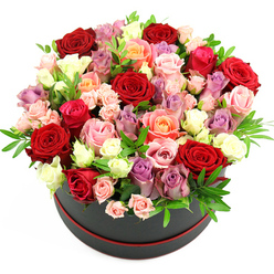 The photograph presents a lavish rose hatbox arrangement, composed with the kind of care and precision you might expect from a seasoned local florist in Brent Cross. The setting is minimal-a clean, white background and slightly elevated viewpoint-so attention falls entirely on the flowers and their elegant container. The hat box itself is a round, matte black cylinder, finished with a slim ring of red trim at the base that acts almost like a subtle frame for the blooms above. From this stylish base emerges a densely packed, dome-shaped bouquet, where roses press closely together in a tapestry of colour. Deep crimson and rich red roses provide a passionate foundation, their petals velvety and tightly layered. Woven between them are blossoming shades of blush pink, vibrant fuchsia, and warm peach, each petal edged softly, creating a gentle gradient of colour as your eye moves across the arrangement. Creamy white and ivory blooms brighten the composition, catching the light and guiding the gaze, while scattered lavender and soft purple roses anchor the piece with a cool, contemporary note. Some roses are fully open, revealing intricate whorls of petals, while others are still cupped or appear as smaller spray roses, giving movement and variety. Soft green foliage, possibly eucalyptus or another fine-leaved filler, peeks through in slender sprigs, breaking up the intensity of the colours and adding a sense of garden freshness. The overall impression is of a premium mixed-rose hatbox gift, ideal for same-day delivery around Brent Cross, whether you're sending love to a partner on an anniversary, congratulating someone on a new home near Hendon, or simply marking a special date with a beautifully composed, romantic gesture.