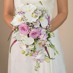 Beneath the soft glow found in Brent Cross, a modern bride holds a breathtaking cascading bouquet that blends timeless romance with contemporary style. The arrangement, engineered with skilled hands, features immaculate white orchids that spill gracefully downward, their silk-soft petals complementing blush pink and lavender roses tucked elegantly within. Baby's breath weaves gently throughout, casting a frothy, ethereal haze that softens the structured lines of the bouquet. Lavender and violet blooms, evocative of summer strolls beside Brent Park, add subtle colour and texture, while trailing magenta amaranthus draws the eye with luxurious, tactile drama. The bride, visible from mid-torso, wears a delicate lace gown; her hands hold the bouquet close, grounding the composition in warmth and anticipation. The muted neutral background focuses attention on the bouquet's detail-each bloom inviting a closer look, each curve reflecting the passion and expertise of Brent Cross Flowers. Altogether, this opulent arrangement brims with the magic of a spring or summer wedding in Brent Cross, from the first flush of new love to the promise of shared tomorrows in North London.