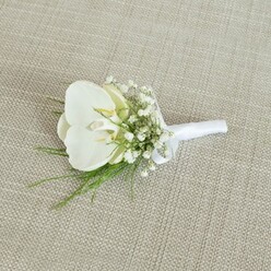 A refined white orchid boutonniere lies gently on a pale beige, woven fabric surface, its subtle linen-like texture giving the scene a natural, tactile warmth. The central flower is a flawless Phalaenopsis orchid, its broad white petals smooth and slightly reflective, catching the soft, diffused light that seems to fall like a calm morning across Brent Cross. At the heart of the orchid, a touch of pale yellow deep in the throat adds a quiet glow, suggesting freshness and the gentle promise of a wedding day just beginning. Surrounding this statement bloom are clusters of white baby's breath, each tiny flower forming part of a delicate, cloud-like ring that softens the edges and adds a romantic haze. Threaded among the gypsophila are wispy strands of bright green foliage, fine and feathery, which add movement and a subtle sense of wildness, as though the boutonniere carries a memory of a walk through a local garden near the River Brent. The stems of the orchid, baby's breath, and greenery are gathered precisely and bound with a smooth white satin ribbon, wrapped in tidy, confident turns, then finished with a small, elegant bow that sits neatly near the base. The neutral background allows the crisp whites and vivid greens to stand out, emphasising the boutonniere's minimalist yet luxurious character. It feels suited to a groom stepping out of a car near Brent Cross station, or a groomsman greeting guests at a North London ceremony, symbolising quiet strength, tenderness, and the care of a skilled florist's hand in every hidden wire and wrap.