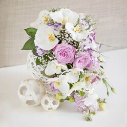A carefully composed floral centerpiece sits on a crisp white surface, conjuring the feel of an elegant wedding table setting in Brent Cross, perhaps moments before guests take their seats. At the heart of the image is a striking white decorative vase shaped like a perfect sphere, its surface formed from slender strands woven together in an airy, open lattice that recalls delicate rattan art. Emerging from this sculptural vessel, an abundant bouquet spreads out in a rounded form, dominated by pristine white Phalaenopsis orchids whose broad, velvety petals catch the light with a gentle sheen. Each orchid displays a refined splash of yellow and gold in the centre, lending the arrangement a quietly luxurious touch. Closely gathered around the orchids are lush roses in soft blush pink and muted lavender, their densely layered petals unfolding in spirals that speak of romance and warmth, ideal for a celebration near Golders Green or a reception close to Hendon Central. Interlaced through the larger blooms, tiny white flowers of baby's breath create a light, cloud-like veil that softens transitions and adds an airy fullness. Ruffled blossoms with white petals tinged in purple and fuchsia tones, likely Lisianthus or Eustoma, peek from between the roses and orchids, adding lively edges of colour and fine detail. From the lower part of the bouquet, a few trailing stems of pale lilac flowers arc downward, suggesting graceful movement and echoing the soft lines of a waterfall bouquet. Fresh green leaves and slender, fern-like foliage provide structure and contrast, deepening the palette without overwhelming the gentle pastels. Resting beside the vase, two cream-toned woven spheres of different sizes mirror the vase's texture and give the arrangement a balanced, thoughtfully curated look. A softly textured, warm beige background remains unobtrusive and calm, ensuring that the flowers, greens, and woven elements stand out as a testament to skillful floristry, the sort you might expect from Brent Cross Flowers for a sophisticated local wedding.