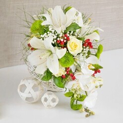 A lavish wedding-style centerpiece stands on a pristine white tabletop, exuding quiet luxury that would feel equally at home in a Brent Cross dining room or hotel banquet suite. The flowers are arranged in a rounded white wicker basket, its tightly woven strands forming a light, textural base that grounds the composition. Rising from this base, large white lilies open wide, their trumpet-shaped blooms edged with soft shadows and decorated with fine, dark speckles, guiding attention to the golden-yellow stamens at the heart of each flower. Near the top-center, a single creamy ivory to pale yellow rose catches the eye, its sculpted petals wrapping gently inwards to form a refined focal point. Flowing gracefully from the right side of the arrangement, white phalaenopsis orchids trail downward, their smooth, rounded petals creating a cascading line that subtly echoes the teardrop shape of a bridal bouquet. Around the main flowers, lush green foliage and textural accents fill the spaces: slender grassy fronds, feathery greens, and bright lime-green pom-pom blooms with a soft, moss-like surface that adds contemporary flair. Tiny white blossoms, reminiscent of baby's breath, peek through the larger blooms, adding an airy veil of delicacy. Deep ruby-red berries are scattered throughout, providing small bursts of colour that enliven the palette, akin to the contrast of seasonal berries against the greenery in Brent Cross's residential gardens. To the left of the main arrangement, two white decorative spheres rest on the tabletop, one large and one small, their open latticework crafted from interlaced strips that echo the basket's design. A soft, neutral beige background with gentle texture completes the scene, focusing attention on the interplay of form, colour, and shadow, and evoking a feeling of joy, purity, and celebration.