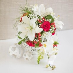 A sophisticated floral arrangement in classic bridal tones of white, red, and fresh green is presented as a centerpiece on a smooth white surface, as if ready for a wedding reception in a Brent Cross hotel or restaurant. The flowers are gathered in a round, open-weave container, its white, basket-like structure made of interlocking strands that form a delicate, airy pattern. Dominating the design are several white oriental lilies, wide open and serene, with gently flared petals and long, pollen-tipped stamens glowing in soft gold. Between them nestle velvety red roses, tightly coiled in elegant spirals, their rich crimson hues adding a dramatic, romantic heartbeat to the arrangement. Sprays of tiny white gypsophila weave among the larger blooms, their small blossoms forming a misty veil that softens every transition. Slender, finely textured greenery threads throughout, echoing the lightness of the gypsophila and giving the piece a sense of movement, like a quiet breeze drifting along the North Circular. Scattered within the flowers, glossy red hypericum berries provide playful bursts of color, reminiscent of festive gatherings or Christmas dinners in the Brent Cross area. Flowing confidently from the right side of the main bouquet, a stem of white Phalaenopsis orchids cascades gently, each bloom with a delicate yellow and blush-pink center and a few buds still closed, promising more beauty to come. On the left of the arrangement, a trio of white woven spheres rest on the table, mirroring the basket's texture and adding a sculptural, contemporary note. The backdrop, a soft, neutral beige with subtle texture, frames the entire design, making it an ideal floral image for wedding décor, engagement parties, or stylish home celebrations around Brent Cross.