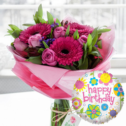 A joyful birthday scene is captured in this image, as if a surprise delivery has just arrived to a home near Brent Cross station on a bright, calm morning. The main focus is a generous, professionally arranged bouquet of fresh flowers set in a clear glass cylinder vase. Dominating the display are large gerbera daisies in vivid shades of fuchsia and magenta, their petals perfectly spaced around dark, almost black centres that lend a rich, velvety depth. Nestled among them are numerous soft pink roses, some tightly furled with promise, others half-open to reveal delicate, layered petals. Between these key blooms, clusters of small, deep purple filler flowers add contrast, with lush, polished green foliage weaving through to create a full, rounded shape reminiscent of hand-tied bouquets often seen in local Brent Cross florists' windows. The stems disappear into a base wrapped in pale, translucent pink cellophane or tissue, crinkled gently and catching the light, giving the arrangement a soft, feminine glow. Around the neck of the vase, a piece of rustic twine is tied into a simple bow and holds a refined off-white gift tag decorated with a fine rose emblem, suggesting a high-quality, carefully curated gift chosen with thought. In the lower right corner, a round foil balloon drifts forward, its glossy surface reflecting the room's light. The balloon's white background is dusted with subtle grey speckles and bordered with stylised flowers and circular motifs in bright yellow, sky blue, lime green, vivid pink, and purple. The words "happy birthday" stretch across the centre in playful pink cursive, echoing the colours of the bouquet. The softly blurred indoor backdrop, likely a Brent Cross home or café with daylight streaming through a window, places all attention on this exuberant floral and balloon pairing, ideal for celebrating a milestone or simple birthday cheer anywhere between Cricklewood and Golders Green.