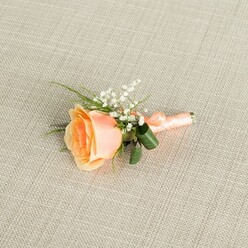 A single peach rose boutonniere is carefully presented on a light beige linen-style fabric, the soft, woven texture giving the image a calm, understated elegance. The rose is the clear focal point, placed slightly off-centre and resting horizontally, with petals that spiral from a warm, sunlit peach core to paler, creamy outer edges. It feels like a bloom cut at just the right moment, still fresh and full of promise. Tucked closely around the rose is a gentle spray of white baby's breath, each tiny flower adding a whisper of softness and a flutter of texture that contrasts with the rose's smooth, velvety petals. Fine, vivid green stems and a couple of darker, broader leaves curve around the base, their glossy surfaces catching the light and giving a hint of movement, as if the foliage has just been shaped in a florist's hand. The stems are wrapped with a satin ribbon in a matching peach shade, wound evenly along the length and secured with a small, neat bow that speaks of meticulous craftsmanship. Soft, diffused lighting glows across the arrangement, highlighting the subtle gradients of colour, the faint sheen of the ribbon, and the even grain of the fabric underneath. The whole boutonniere suggests a moment of poised anticipation, ideal for a groom stepping out of a townhouse near Brent Cross or a prom guest arriving from nearby Hendon or Cricklewood. It has the intimate charm of a bespoke piece made to order for a local celebration, ready to be pinned to a lapel for a wedding, engagement party, or formal dinner in the Brent Cross area.