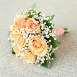 This bouquet, as envisioned by a skilled Brent Cross florist, radiates a gentle sophistication befitting special occasions held near Clitterhouse Playing Fields or within the lively residential heart of NW4. The core of the composition is a handful of luxuriant roses tinged in shades of peach, apricot, and pale sunrise, their petals coiling in elegant spirals and glowing softly in the subtle daylight. Feathery Gypsophila-known as baby's breath-envelops the roses in dreamy, cloud-like clusters, their bright whiteness heightening the warmth of the roses and infusing the bouquet with a note of airy lightness. Hints of glossy emerald foliage peek from between the blossoms, adding natural definition and a freshness reminiscent of local gardens in full bloom. Each flower stem is hand-gathered into a neat, classic dome and secured with a blush-toned satin ribbon, its luxurious texture enhanced by the gentle spiral of small white faux pearls that wrap the handle-an elegant nod to traditional bridal accessories often seen at Brent Cross celebrations. The exposed, clean-cut stems hint at the garden-to-hand freshness of every arrangement. Resting on a textured fabric canvas, this bouquet is subtly bathed in soft, natural light, highlighting every gentle curve, petal edge, and shimmering pearl. Designed for bridesmaids, romantic anniversaries, or heartfelt milestones across Brent Cross and Hendon, it invites every recipient to experience its lasting beauty and tenderness.