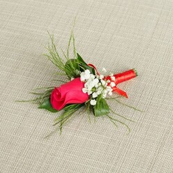 On a pale, sand-toned fabric background with a subtle woven texture, a delicately made boutonniere is positioned at a soft diagonal, as if just placed down by a careful hand before being pinned onto a jacket in Brent Cross. The focal point is a single rosebud in a vivid fuchsia-pink, its petals tightly wrapped and smooth, like satin folded in upon itself, holding a sense of anticipation that suits a wedding morning or an anniversary celebration. The rose's rich colour deepens toward the center, moving from bright pink to a more intense magenta, and is illuminated by bright, natural-style light that highlights every curve of the petals. Encircling this central bloom are tiny white baby's breath flowers, each cluster forming a misty, cloud-like ring that softens the intense hue of the rose and adds texture without overwhelming it. Fresh green foliage frames the arrangement: some sprigs are fine and feathery, slightly arching outward, while a few slightly broader leaves provide a quiet, grounding base. Together, the greenery creates a feeling of lightness and movement, as if a gentle breeze from nearby Clitterhouse Playing Fields had passed through. At the base, the slender stems are bound with a coral-red satin ribbon, carefully wound in smooth, even coils that gleam softly where the light catches them. The ribbon is secured neatly, suggesting the practiced touch of a local florist used to preparing buttonholes for Brent Cross weddings and school proms across nearby neighbourhoods like Golders Green and Cricklewood. The overall mood is one of tender elegance and heartfelt sentiment, a small but meaningful floral detail that can accompany shared vows, first dances, and cherished family photographs.