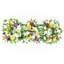 The image shows a beautifully balanced funeral flower tribute spelling "DAD," set against a clean, bright white background that highlights the rich colours and thoughtful detailing. Each of the three capital letters - the first D, the central A, and the final D - stands side by side in a neat row, their forms softened by a collar of dark green fern foliage. The fern fronds curve gently around the edges, introducing a sense of natural movement and echoing the greenery found in parks and residential gardens near Brent Cross. Within this green frame, each letter is tightly filled with velvety white chrysanthemums and open white gerbera daisies, their petals overlapping to create a plush, cushion-like surface symbolising purity, reverence, and cherished memories. Scattered throughout the white base are bright yellow gerbera daisies and tiny yellow spray roses or button blooms, adding warm, sunlit notes that feel like flashes of happy family moments. Adding a deeper emotional tone, sprigs of purple lisianthus and clusters of magenta or deep pink spray roses are placed thoughtfully among the white and yellow, with small lavender-hued filler flowers and bright green button chrysanthemums tucked between them for extra texture and contrast. The arrangement is entirely front-facing so that the word "DAD" reads clearly, making it ideal as a central tribute at a church, crematorium, or graveside service around Brent Cross and the surrounding area. Slim, dark support bars quietly join the letters from behind, ensuring they remain straight and unified while remaining unobtrusive. The overall design feels like a heartfelt, bespoke tribute crafted by Brent Cross Flowers, created to honour a father's life with elegance, warmth, and sincere affection.