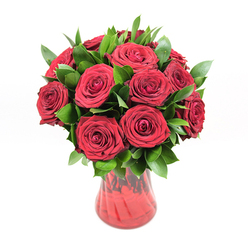 A rich, romantic bouquet of deep crimson red roses stands proudly in a translucent red glass vase, photographed against a bright white background that makes every colour seem more vivid. There are roughly twelve to fifteen roses, arranged in a full, rounded dome, each bloom opened to reveal layers of velvety petals curving inward in perfect spirals. The red is intense and luxurious, like garnet silk, and the slightly ruffled edges of the petals create soft shadows that lend the arrangement real depth. Threaded throughout the flowers is a generous amount of dark green foliage, probably ruscus, its smooth, lance-shaped leaves emerging between the blossoms to frame them and give the bouquet a lush, garden-fresh look. The leaves appear glossy under the lighting, a cool counterpoint to the warmth of the red. The vase rises in a gentle curve, narrowing toward the base and then flaring at the top, its semi-transparent red glass showing subtle ribbing or wave-like texture that catches reflections. You can just make out the stems inside, blurred by the glass, suggesting the honest structure behind the polished design. The clean, uncluttered white backdrop throws all attention onto the interplay of red and green, making this design feel both contemporary and timeless. It's easy to imagine this arrangement being hand-delivered to a home near Brent Cross Shopping Centre or a quiet street by Hendon Park, a passionate gesture for an anniversary, romantic birthday, or intimate celebration. The bouquet seems to radiate devotion and enduring love, the sort of rose arrangement chosen when words feel too small.
