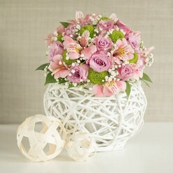 Imagine the gentle, uplifting mood as this floral arrangement graces a Brent Cross dining table, setting the stage for family celebration or a romantic anniversary. The plush centerpiece abounds with pastel pink and soft lavender roses, their velvet petals gently unfurling to catch the diffused daylight-a testament to the freshness and artistry at Brent Cross Flowers. Alstroemeria, with their slender petals boasting graceful stripes and occasional splashes of yellow, mingle with the roses, channeling the area's vibrant character. Pops of lime green from well-placed button chrysanthemums invigorate the bouquet, much like a sudden sunbeam across the green open spaces off Hendon Way. Wisps of tiny white baby's breath thread through the composition, lending a gauzy airiness that softens the overall look, while scattered dark green foliage grounds it in the region's familiar leafy abundance. Everything is unified in a showpiece white vase-its distinctive, open-woven architecture blending modern design with a touch of rustic charm, reminiscent of handwoven crafts sourced from local Brent Cross artisans. To the left, two decorative spheres in matching woven texture-one slightly larger than the other-echo the vase's motifs and add whimsical visual rhythm. The softly textured light beige wall behind ensures every colour and petal takes centre stage. Altogether, this image embodies artisanal pride, grace, and celebratory spirit, perfectly suited to wedding settings or as a sophisticated accent for a Brent Cross home.