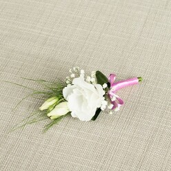 An elegant floral buttonhole lies diagonally across a pale beige, linen-like fabric, the subtle weave of the background giving the scene a gentle, natural texture reminiscent of a well-set table in a Brent Cross townhouse. The centerpiece of the arrangement is a pristine white lisianthus bloom, fully open and generously ruffled, its overlapping petals creating a soft, almost cloud-like cup of white that suggests purity, calm, and understated luxury. Nestled just to the left and slightly below are two elongated eustoma buds, still closed and tinted with soft cream and hints of fresh green, echoing the promise and anticipation of a wedding morning. Threaded delicately throughout are clusters of tiny white baby's breath, each miniature blossom dotting the boutonniere with a lacy, starry quality that enhances the sense of lightness and romance. Behind the flowers, several slender, grass-like strands of greenery arc outwards, their smooth, linear forms adding movement and contrast to the rounded petals, while a single, broader deep green leaf acts as a quiet backdrop, giving depth and definition. The gathered stems are tightly wrapped in a striking fuchsia satin ribbon, tied into a small, confident bow at the base, its bright pink tone injecting a playful, modern accent against the otherwise classic palette. The satin catches the soft light, creating gentle reflections that draw the eye down the length of the stems. The composition feels intimate and thoughtfully balanced, perfect for a groom or groomsman stepping out of a car near Brent Cross Shopping Centre or walking through nearby leafy streets, carrying just a touch of floral grace and celebration on their lapel.