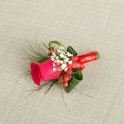 A carefully crafted floral buttonhole lies horizontally across a pale beige, subtly woven fabric, reminiscent of a linen jacket laid out before a ceremony in Brent Cross. The central flower is a deep fuchsia rose, its petals lush and tightly layered, forming a rich spiral that suggests both energy and tenderness. Soft daylight glides across the bloom, catching the edges of the petals so that the pink shifts from almost magenta at the centre to a gentler rose tone near the outer edges. Clustered neatly at the base of this focal flower are sprigs of white baby's breath, each cluster made up of dozens of pinhead blossoms, giving the arrangement an airy, cloudlike halo. Dotted between them, plump red hypericum berries bring a lively contrast, their polished skins glowing against the delicacy of the gypsophila. A mix of greenery surrounds and supports the design: fine, feathery fronds stretch lightly outward, while darker, broader leaves are artfully folded and curled to cradle the rose, adding depth and structure. The stems are bound with a smooth, rich red satin ribbon, carefully wound and finished in a compact bow that signals both elegance and comfort for the wearer. The boutonniere is photographed from a slightly elevated angle, so every detail-the curve of each petal, the berries' roundness, the gentle sheen of the ribbon-can be appreciated. It feels like a small but meaningful token, perfect for a groom or groomsman stepping out onto the streets around Tilling Road or heading to a reception near Brent Cross Shopping Centre, carrying a touch of local floral artistry close to the heart.