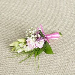 A refined boutonniere or petite corsage lies perfectly still on a pale, woven fabric that looks like fine linen, its warm beige tones echoing the calm, elegant interiors so often seen in Brent Cross townhouse reception rooms. The floral work is compact but full of character: at its heart, a cluster of soft pink, ruffled flowers, close in style to miniature stocks or romantic garden blooms, gathers tightly together, each petal curling and overlapping to create a plush, cushion-like texture. Tiny white baby's breath flowers are interspersed throughout, their cloud-light blossoms adding a lace-like delicacy that softens the arrangement. Flickers of lavender or deep lilac florets nestle between the pink and white, offering a gentle hint of richness and depth, like dusk shadows along the nearby Hendon Way. On one side, several slender blooms in creamy white and pale green remain closed, their elongated buds evoking freesias just on the verge of opening, capturing that quiet sense of anticipation that often surrounds a wedding morning. The whole design sits upon a base of lush, emerald green leaves, their broad surfaces smooth and slightly reflective, while fine, airy sprigs of fern-like greenery and grass-thin fronds extend beyond the edges, introducing a naturally untamed finish. At the base, the neatly aligned stems are wrapped in a vivid fuchsia satin ribbon, the bow tied with a florist's practiced hand so that its loops are balanced and its tails fall neatly across the fabric. The gentle, even lighting highlights every texture-from the soft petals to the cool, sleek ribbon-making this piece an ideal finishing touch for a groom, bridesmaid, or prom guest heading out from Brent Cross for an evening celebration.