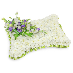 This rectangular floral pillow reads like a tactile embrace: hundreds of small white chrysanthemum heads are set tightly together to form a uniformly soft surface, their layered petals creating a serene, cloud-like texture that symbolizes purity and rest. The pillow is bordered all the way around with a light lime-green ribbon, hand-pleated into neat overlapping loops that provide a crisp, gentle frame and a quiet note of hope. Diagonally placed in the upper left is a composed cluster of accent blooms - crisp white lisianthus paired with rich, deep purple lisianthus and a few upward-closed purple buds - which give the composition a respectful counterpoint of colour and depth. Wisps of feathery fern fronds and broader green leaves are woven through the cluster, introducing natural movement and soft contrast, and a dark green leaf wrap at the base keeps the stems tidy and refined. The pillow's slightly raised profile and subtle shadow on the studio-white surface communicate careful craftsmanship and dimensional weight, as if it could be gently rested upon. The scent is delicate and reassuring, the cool green foliage tempering the chrysanthemum's soft floral tone. Designed as a dignified sympathy piece for funerals, memorial services, and gravesides, it expresses condolence in a quiet, traditional language. Locally, this style is often chosen for services around Brent Cross and neighbouring Hendon or Golders Green chapels, where families look for arrangements that speak of calm, respect, and enduring remembrance.