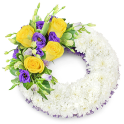 A pristine round sympathy wreath pictured on a plain white backdrop, its substantial form created from countless tight white spray chrysanthemums that give the piece a cloud-like, reverent surface. The dense white base feels tactile and uniform, an intentionally serene field upon which a colourful spray rests at the upper left: rich yellow roses, their petals unfurling in warm spirals, nestle against luxuriant, deep purple lisianthus with elegant ruffled edges. Small lavender and pale green lisianthus buds, along with slender fern fronds and glossy, slightly variegated foliage, add layered contrast and a measured sense of life within the arrangement. Delicate sprigs of tiny white fillers and a faint hint of dark purple crinoline or mesh ribbon showing at the wreath's inner and outer rims subtly tie the accent colours back to the foundation. The open centre reinforces the traditional wreath silhouette while soft morning light highlights the roses' velvety warmth and the chrysanthemums' soft sheen. From a florist's perspective familiar with Brent Cross - passing the shopping centre lanes or arranging tributes for services near the Welsh Harp - this composition reads as both dignified and comforting. Brent Cross Flowers crafts this style of tribute for local memorials; the design is deliberately restrained, offering mourners a message of peace, memory and gentle farewell rendered with careful technique and seasonal freshness.