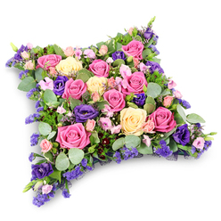 In this image, a beautifully composed funeral cushion takes centre stage, a floral pillow tribute thoughtfully designed in a soft-edged rectangular shape that suggests comfort and rest. Every inch of the cushion's surface is crowded with flowers, so that only petals and foliage meet the eye. Roses in several shades are the stars: vivid fuchsia blooms bring a lively yet tender energy, while softer blush pink and creamy pale yellow roses add light and gentleness, their velvety petals overlapping in layered whorls. Among these, royal purple lisianthus flowers weave through the design, their ruffled, cup-shaped heads adding a luxurious contrast that deepens the colour story. Fine sprays of smaller pink roses and delicate clusters of tiny lavender-toned filler blossoms, akin to statice or limonium, fill even the most hidden gaps, giving the cushion a carefully finished, almost embroidered appearance. The edges and lower layers are softened with an array of greenery: rounded, silvery eucalyptus leaves catch the light with a muted gleam, while darker, fern-like foliage brings an airy, textural frame. Set against a pure white background, the colours feel especially vivid yet remain refined, creating a mood that is both serene and heartfelt. The arrangement seems ideal for a funeral or memorial service in and around Brent Cross, a floral tribute that might rest beside a coffin in a synagogue or chapel, or be placed lovingly at a graveside. It embodies the gentle message that, even in loss, there is beauty, care, and a wish for peace, lovingly conveyed through expertly chosen blooms and considered design.