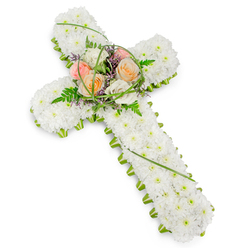 A traditional Christian cross made entirely from fresh flowers is pictured against a pristine white background, giving the arrangement a sense of calm and focus, as if it were placed in a quiet chapel near Brent Cross just before a service begins. The body of the cross is densely massed with small white chrysanthemums, each head nestled tightly against the next to form a continuous, velvety carpet of petals. This unbroken white surface suggests purity, reverence, and a soft place for the eye and heart to rest. Tracing the perimeter of the cross, a slim border of bright green foliage adds structure and clarity, accentuating the outline in much the same way that tree-lined pathways define the edges of local parks and gardens around Brent Cross and Golders Green. At the very centre, where the vertical and horizontal sections meet, a prominent spray of colourful yet gentle blooms becomes the focal point. Several roses in warm peach, tender blush, and creamy ivory tones open gracefully, their layered petals curling outward in soft spirals. These roses are carefully interwoven with delicate fern fronds, polished green leaves, and arching strands of narrow, bright green grass that bring movement and a sense of renewal. Tiny flecks of purple or lavender filler flowers are sprinkled throughout, providing subtle contrast and depth. The cross is photographed on a slight angle, letting the viewer follow the line of flowers from top to base. This arrangement feels especially fitting for a funeral, memorial, or graveside ceremony across Brent Cross, speaking quietly of faith, hope, and the enduring bond with a loved one who has passed.