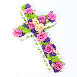 Seen from above against a pure white background, this substantial floral cross appears almost to float, its diagonal positioning giving movement and presence. The outline is shaped by a thick border of white spray chrysanthemums, their small, neat petals forming a continuous, cushiony edge that softens the geometry of the cross. Just beneath this white border, a band of light-green ribbon or closely wrapped foliage encircles the design, its subtle pleats adding both structure and a fresh, verdant accent. Inside this frame, the interior of the cross is alive with colour. Generous roses in radiant hot pink and creamy pale yellow, some softened with a hint of peach, are placed in clusters along the arms and down the centre, their open blooms revealing intricate spirals of velvety petals. Scattered among them are vivid lime-green button chrysanthemums, each bloom a small, rounded pom-pom that introduces cheerful texture and contrast. Woven around and between these focal flowers are smaller, deep purple and violet blossoms-likely lisianthus or statice-bringing a dusk-like depth to the arrangement, much like the cool shadows cast along the paths near Brent Cross. The lighting is bright and even, with no harsh shadows, so each petal edge, each gleam of ribbon, stands out clearly against the uncluttered background. This floral cross is unmistakably a funeral or sympathy tribute, traditional in its Christian shape yet modern in its lively colour palette. It is the sort of arrangement that might be chosen by families around Brent Cross to convey both solemn remembrance and a gentle sense of hope, whether placed by a church altar, at a crematorium chapel, or laid tenderly at a graveside.