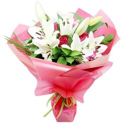 In this thoughtfully composed bouquet, tall white Oriental lilies and tender pink roses are gathered into a lush, hand-tied arrangement, shown clearly against a bright white background. The lilies, some fully open and others still in elegant bud form, form the sculptural backbone of the design. Their pure white petals flare outward in trumpet-like shapes, while at the heart of each bloom, deep orange-brown anthers sit atop slender filaments, a subtle dusting of pollen visible on some petals. The unopened buds, elongated and edged with soft green, suggest that the bouquet will continue to evolve over several days, rewarding the recipient with fresh blooms. Nestled below and between the lilies, smaller pink roses provide a gentle, romantic counterpoint. Their petals, tightly coiled and slightly satiny, range from pale blush to a warmer rose-pink tone as they spiral inward, adding both colour and texture. Around this floral core, a generous collar of foliage spreads outward, with rich green leaves in varied shapes and sizes lending depth and a natural garden feel, not unlike the greenery you might see along quiet residential streets around Brent Cross or in nearby parks. The entire bouquet is wrapped in generous layers of shiny pink cellophane or floral paper, arranged in soft ruffles and folds that catch reflections and frame the flowers like a celebratory halo. At the base, a slim lime green ribbon is tied carefully around the stems, creating a fresh contrast and giving the arrangement a polished finish. This bouquet would make a beautiful delivery to a Brent Cross home or office, ideal for marking a new job, a special anniversary, or simply sending a message of admiration and hope.
