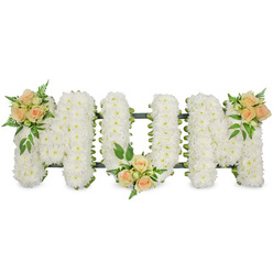 This image shows a beautifully composed "MOM" funeral tribute laid out horizontally, the three capital letters standing out crisply against a seamless white background as though floating in soft light. Each letter is formed on an unseen framework and completely blanketed with small, delicate white blossoms-most likely spray chrysanthemums or similar blooms-packed so closely together that they create a thick, cushiony texture. The petals overlap in layers, giving a velvety, cloud-like effect that conveys purity and calm, just the sort of arrangement that might be placed beside a coffin in a Brent Cross funeral home or delivered to a chapel near the shopping centre. Around the outer edges of the letters, a faint line of dark green frame and ribbon is visible, providing just enough contrast to define the graceful shapes without distracting from the flowers themselves.   Soft colour is introduced through three floral focal points of peach and apricot spray roses. One cluster of roses decorates the upper left arm of the first "M", the second rests gently at the base of the central "O", and the third graces the upper right arm of the final "M", giving a sense of balance and rhythm across the tribute. The roses vary from tight buds to half-open and fully open blooms, adding visual interest and a feeling of life and unfolding stories. Around each group of roses, small tufts of greenery-light, ferny textures mixed with firmer, darker leaves-spill out slightly over the bed of white flowers, bringing a soft, natural feel. The overall impression is of a lovingly crafted, personal farewell, perfect for families across Brent Cross, Cricklewood, and Finchley who wish to honour a devoted mother with grace and gentleness.