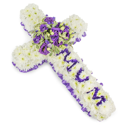This solemn, thoughtfully arranged floral cross is built from a solid mattress of bright white chrysanthemum blooms, their rounded petals creating a cloud-like surface that reads as both tender and steadfast. The image captures the tribute on a clear white studio background so every detail is visible: a scalloped, deep purple satin ribbon traces the outer edge, its ruffled texture forming a composed border that contrasts with the softness of the chrysanthemums. Running down the upright shaft, the word "MUM" appears in clearly defined purple letters made from small lisianthus or compact spray chrysanthemums, the letters raised slightly so they stand out against the white field. At the centre a rich focal spray brings the arrangement to life-a compact bouquet of deep violet roses and lighter lavender lisianthus, supported by variegated foliage with pale margins and darker centres, along with tiny green berries that punctuate the cluster and add a subtle, living accent. The palette of snowy white and layered purples suggests a mixture of peace, devotion and dignified sorrow; the design reads as both classic and quietly personal. You can imagine the gentle, powdery scent of chrysanthemums mingling with the sweeter, tea-like perfume of the roses. As an experienced florist in Brent Cross I assemble pieces like this with attention to stability for graveside laying and chapel placement; Brent Cross Flowers can prepare such a tribute for services across Hendon, Cricklewood Broadway and the surrounding neighbourhoods, honouring memory with careful craftsmanship and calm presence.