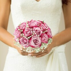 In the gentle morning light of Brent Cross, this close-up captures a bride adorning her wedding ensemble with a bouquet that seems to breathe elegance. Roughly a dozen roses, each flawlessly shaped, create a harmony of pastel and fuchsia hues-their petals unfolding from tender pink at the core to boldly vibrant edges. Wisps of baby's breath, luminous and white, drift among the roses, lending an ethereal, almost cloud-like quality that softens the intensity of the pinks. The bride's hands, radiating a warm skin tone beneath an artful, light manicure, hold the bouquet lovingly against the refined, lace-adorned bodice of her gown. The sleeveless dress, dreamy in its design with a V neckline and a graceful sash detail, whispers of classic Brent Cross style-blending tradition with understated glamour, echoing scenes from nearby Temple Fortune and local celebratory halls. The soft, neutral backdrop gently fades, drawing all eyes to the bouquet's artistry. This arrangement encapsulates the quiet excitement of a wedding day, the bouquet an emblem of love's promise, expertly crafted for moments destined to be remembered and shared in North West London.