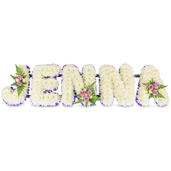 This memorial arrangement gently spells JENNA in sculpted, capital letters formed from dense, creamy-white chrysanthemums that sit like small pillows of bloom. Each letter's softness is contrasted by a neat ribbon of deep lavender-purple outlining its inner edges, lending definition and a touch of refined color without disturbing the serene white field. Three small, hand-tied clusters punctuate the composition-one on the J's curve, another on the middle of the second N, and a final cluster on the A-composed of blush-pink and pale lilac spray roses accented with a few light cream roses and delicate fern fronds. The roses add warmth and a whisper of scent; the ferns bring a cool, textured green that moves the eye across the name. Thin, dark metal support bars are discreetly visible, connecting the letters and ensuring the tribute remains stable whether displayed at a wake or placed by a graveside. The overall effect is both tender and dignified: a personalized floral message that reads clearly from a distance and invites quiet reflection up close. Light falls softly across the arrangement, throwing gentle shadows in the grooves between letters and making the ribboned edges glow slightly, as if lit for an intimate service. Created with local insight into Brent Cross and nearby Hendon funeral venues, this piece feels handcrafted and thoughtfully composed-an elegant, compassionate way to honour a life and name with floral grace.