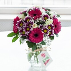 A richly coloured, professionally arranged bouquet sits centre stage in a clear, textured glass vase, bathed in soft natural light that suggests a bright Brent Cross morning. The flowers form a rounded, abundant dome of colour, anchored by large gerbera daisies in deep fuchsia and magenta shades, their dark, velvety centres adding drama and depth. Around them, generously petalled chrysanthemums in striking purple-and-white bicolour create a lively ring of texture; each blossom has purple-edged petals that gradually soften to white around a sunshine-yellow centre, reminiscent of late-summer garden borders in nearby Hendon. Threaded gently through the bolder blooms are clusters of creamy white spray roses, some fully open with layered, satiny petals, others held in tight buds that promise to unfurl over the coming days. Wisps of delicate white baby's breath (gypsophila) float between the larger heads, giving the arrangement a soft, cloud-like quality and enhancing the sense of airiness. Deep green foliage frames the arrangement, its cool tones balancing the warm pinks and purples and adding a natural, garden-fresh feel. The short, jar-style vase is wrapped at the neck with a simple loop of rustic twine tied in a neat bow, from which hangs a small white tag bearing a stylised pink rose motif and elegant script, as though ready for a personalised message from a local sender. The softly blurred, white background keeps all attention on the vibrant flowers, suggesting this could be a freshly delivered bouquet set on a windowsill in a home off the North Circular, bringing colour, celebration, and an uplifting scent to an ordinary weekday in NW11.
