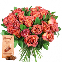 As a florist rooted in Brent Cross, I see this luxurious set as a heartfelt expression of color, joy, and community spirit, perfect for those cherished moments along Edgware Road or in the shadow of Brent Reservoir. The abundant bouquet is a radiant collection of roses in shades that evoke a painter's palette-gentle creams and pale yellows at their centres, expanding outward into a glowing spectrum of peach, coral, salmon, and vivid pink. The petals are silk-soft, unfurling softly, each one illuminated by the clean white background as though touched by morning sun over West Hendon. Rich emerald foliage frames the blooms, offering a glossy, textural border that amplifies their vibrancy and suggests a sense of movement, much like the leafy pathways of nearby parks. Complementing the flowers is a light beige Thorntons Orange Crisp chocolates pouch, its elegant design and illustration promising a decadent treat-a tangy orange truffle with shards of crispy wafer, all enveloped in milk chocolate. Two sleek, unwrapped truffles lie before the pouch, hinting at silky sweetness and citrus fragrance. This thoughtfully composed set, available for delivery throughout Brent Cross and surrounding communities, is ideal for celebrating milestones, comforting a friend, or simply sharing delight. It reflects not only the art of floristry but also the welcoming heart of North West London's neighborhoods.