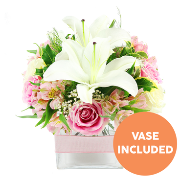 This image presents a carefully composed bouquet set against a pristine white background, the simplicity of the setting allowing every flower to stand out as if lit by soft daylight filtering into a Brent Cross apartment. At the centre of the picture, two fully open white Oriental lilies form the main focal point, their sculptural petals gently arching back to frame clusters of bold reddish-brown stamens and central pistils. Encircling the lilies, soft pink roses form a rounded collar, with petals in various stages-some still tightly wound in a classic spiral and others relaxed and open, revealing layer upon layer of smooth, velvety texture that speaks of romance and gentle celebration. Interspersed among the larger blooms are stems of pink and white alstroemeria, their elegant petals slightly flared, several lightly speckled with yellow, adding subtle pattern and interest to the design. Wisps of delicate white baby's breath weave through the bouquet like tiny stars, lending a sense of lightness and filling the spaces without overwhelming the central flowers. The arrangement is backed by rich greenery, including broad, shiny leaves that reflect the light and feathery fronds that introduce a sense of movement, similar to trees swaying along the paths near Brent Cross Park. All the stems converge neatly in a low, square clear glass vase, the waterline visible and the green stems clearly seen, giving the design a fresh, contemporary feel. Around the middle of the vase, a pale pink satin ribbon is wrapped smoothly, adding a soft decorative accent. In the lower right corner of the image, an orange circular badge with bold white capital text reads VASE INCLUDED, clearly highlighting that the vase forms part of the floral gift, ideal for same-day flower delivery to homes and offices around Tilling Road and the North Circular.
