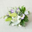 A hand-tied bouquet of white lilies and purple lisianthus lies diagonally across a soft, light beige fabric, as if just set down on a dressing table in a Brent Cross town house before a celebration. The lilies are the stars of the design: large, velvety white blooms fully open, their petals curving gracefully back to reveal warm yellow stamens dusted with pollen and faint cinnamon-speckled throats. Around them, several fresh chartreuse-green buds point outward, promising more flowers in the days ahead and adding a sense of movement and growth. Threaded between the lilies are ruffled lisianthus in creamy white trimmed with vivid purple and violet, their frilled petals forming little cups that echo the gentle folds of a silk gown. Tiny heads of white gypsophila float in between like flecks of foam, softening the lines and giving the bouquet a cloud-like lightness. Lush greenery cradles the blooms, a mix of broad, glossy dark leaves and finer, more feathery fronds that stretch and arc from the edges, suggesting a garden just beyond Hendon Way or near Brent Cross Shopping Centre. The stems are carefully gathered and wrapped in a smooth white satin ribbon, dotted delicately with pearl-like beads that glint subtly where the light touches them, reinforcing the idea of a bridal bouquet, bridesmaid flowers, or a special presentation piece. The textured beige backdrop is calm and understated, allowing the white, green, and purple palette to truly sing, while the soft, diffused lighting makes the whole arrangement feel freshly made, as though prepared that morning by an attentive local florist ready to deliver across Brent Cross for a wedding, anniversary, or intimate family gathering.